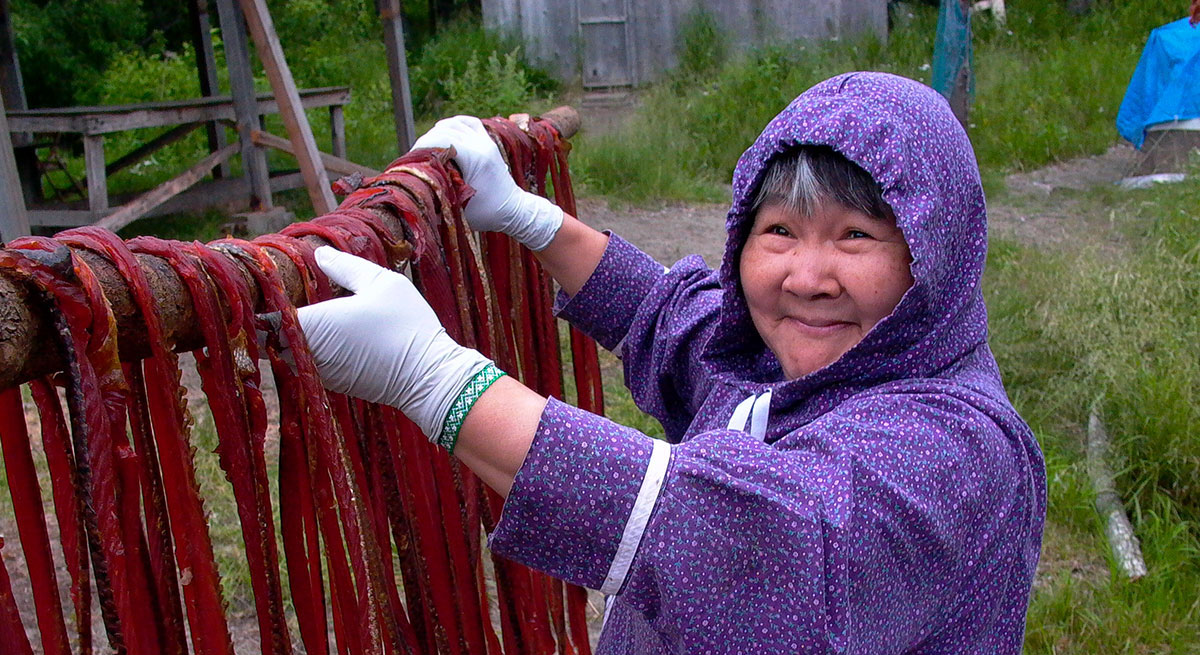A smiling woman in a purple flower-print coat, drying fish on a rack outdoors