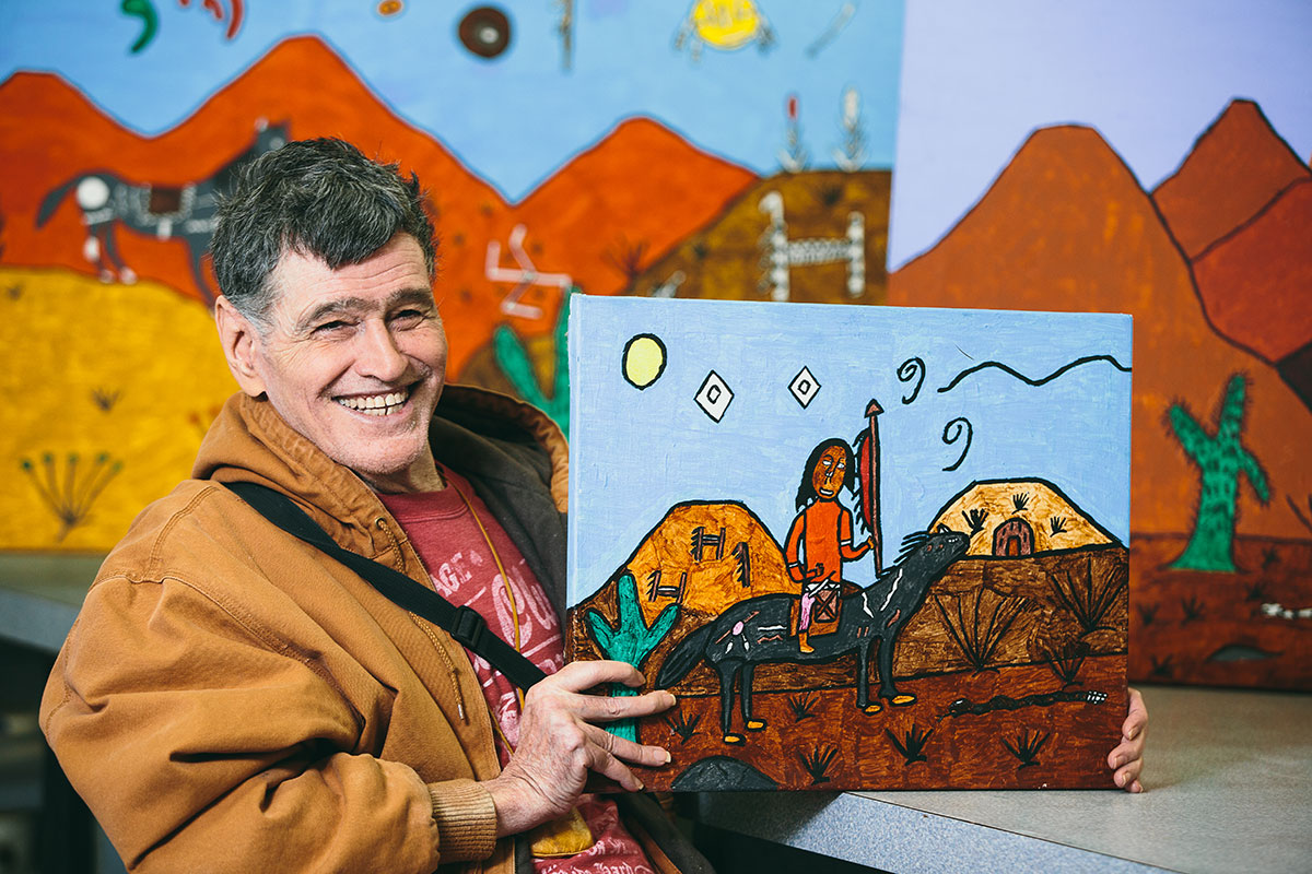 A smiling man holding up a painting, sitting in front of a larger painting done in the same style